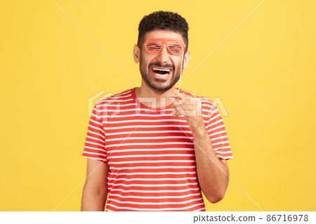 Extremely happy excited man with beard in striped t-shirt holding mask on stick hear eyes pretending he is in eyeglasses, having fun Extremely happy excited man with beard in striped t-shirt holding mask on stick hear eyes pretending he is in eyeglasses, having fun 86716978