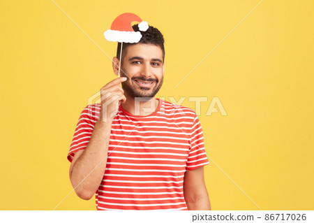 Positive smiling man with beard in red striped t-shirt holding mask on stick with santa claus hat, preparing for christmas and new year celebration. Indoor studio shot isolated on yellow background Positive smiling man with beard in red striped t-shirt holding mask on stick with santa claus hat, preparing for christmas and new year celebration. Indoor studio shot isolated on yellow background 86717026