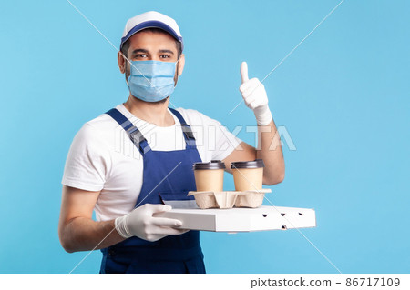 Delivery service. Friendly optimistic courier in overalls and mask holding coffee and pizza box, wearing safety gloves offering drinks food and showing thumbs up, like gesture. indoor, isolated 86717109