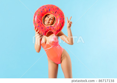 Portrait of cheerful pretty woman in swimsuit looking through big donut rubber ring and showing victory gesture, having rest on summer beach, vacation resort. studio shot isolated on blue background Portrait of cheerful pretty woman in swimsuit looking through big donut rubber ring and showing victory gesture, having rest on summer beach, vacation resort. studio shot isolated on blue background 86717138