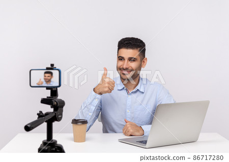Online webinar, distant training. Elegant successful businessman sitting at workplace, showing thumbs up to mobile phone while recording course, doing blog. studio shot isolated on white background Online webinar, distant training. Elegant successful businessman sitting at workplace, showing thumbs up to mobile phone while recording course, doing blog. studio shot isolated on white background 86717280