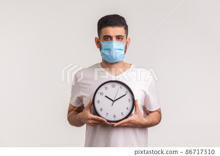 Time to stop coronavirus. Man in protective hygienic mask holding clock, warning of novel virus epidemic, quarantine and risk during incubation period. indoor studio shot isolated on white background Time to stop coronavirus. Man in protective hygienic mask holding clock, warning of novel virus epidemic, quarantine and risk during incubation period. indoor studio shot isolated on white background 86717310