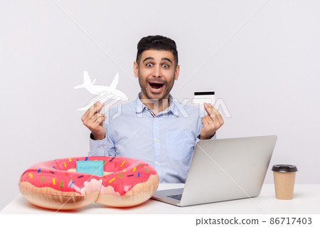 Amazed shocked man holding paper airplane and bank card, sitting in office workplace with rubber ring passport on desk, booking tickets, buying tour, weekend trip. studio shot isolated, travel concept Amazed shocked man holding paper airplane and bank card, sitting in office workplace with rubber ring passport on desk, booking tickets, buying tour, weekend trip. studio shot isolated, travel concept 86717403