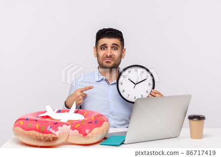 Time to rest. Upset man showing clock, looking frustrated tired at camera, sitting in office workplace with rubber ring passport and paper airplane on desk, employee needs relax travel. studio shot Time to rest. Upset man showing clock, looking frustrated tired at camera, sitting in office workplace with rubber ring passport and paper airplane on desk, employee needs relax travel. studio shot 86717419