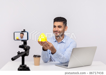 Positive smiling man, real estate agent sitting in office workplace and showing paper house to camera, recording video, streaming online blog about purchase or rental housing. studio shot isolated Positive smiling man, real estate agent sitting in office workplace and showing paper house to camera, recording video, streaming online blog about purchase or rental housing. studio shot isolated 86717438