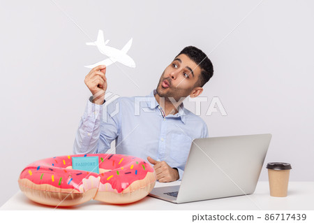 Joyful carefree man playing with paper airplane, sitting in office workplace with rubber ring passport on desk, dreaming of travel vacation, weekend trip. studio shot isolated on white background Joyful carefree man playing with paper airplane, sitting in office workplace with rubber ring passport on desk, dreaming of travel vacation, weekend trip. studio shot isolated on white background 86717439