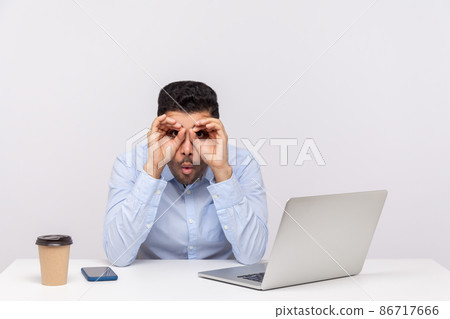 Shocked man employee sitting office workplace with laptop on desk, looking through binoculars hand gesture and expressing big surprise amazement. indoor studio shot isolated on white background Shocked man employee sitting office workplace with laptop on desk, looking through binoculars hand gesture and expressing big surprise amazement. indoor studio shot isolated on white background 86717666