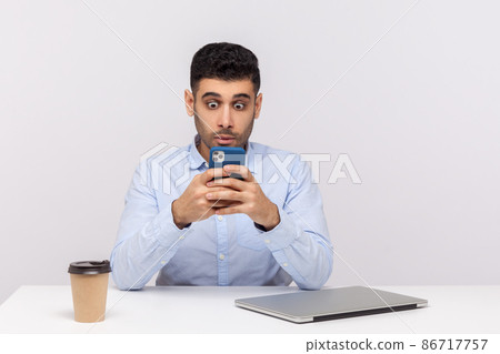 Amazed businessman sitting in office workplace reading shocking message on cellphone, using mobile application with surprised astonished expression. indoor studio shot isolated on white background Amazed businessman sitting in office workplace reading shocking message on cellphone, using mobile application with surprised astonished expression. indoor studio shot isolated on white background 86717757