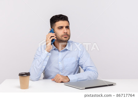 Serious worried businessman sitting in office workplace with laptop, talking on mobile phone, calling partner to consult, discussing bad news, negotiations. studio shot isolated on white background 86717764