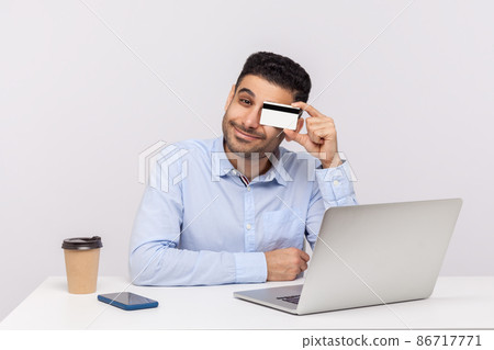 Positive playful man sitting at workplace with laptop, covering eye with credit card and smiling. E-commerce and bank services, online purchase concept. indoor studio shot isolated on white background 86717771