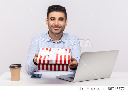 Positive man employee sitting in office workplace, holding gift box and smiling friendly, giving present to camera, congratulations on professional holiday. studio shot isolated on white background 86717772