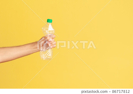 Profile portrait male hand holding and pressing plastic bottle with green cap, sorting his rubbish, worrying about environmental pollution. Indoor studio shot isolated on yellow background 86717812