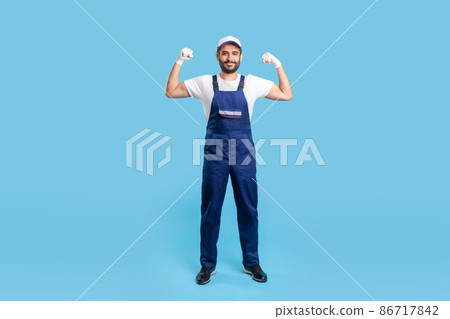 Full length cheerful handyman in overalls, cap and protective gloves standing, showing biceps, demonstrating strength. Profession of service industry, loader or building worker. studio shot isolated 86717842
