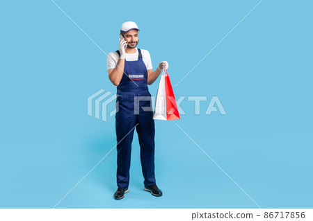 Full length portrait of smiling courier in workwear talking on cellphone, holding shopping bags and communicating with client. Delivery man answering phone call, accepting order at fashion store Full length portrait of smiling courier in workwear talking on cellphone, holding shopping bags and communicating with client. Delivery man answering phone call, accepting order at fashion store 86717856