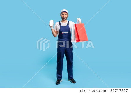 Full length positive professional courier in uniform holding shopping bags and mobile phone with mock up blank display for advertise image, online delivery order at fashion store. studio shot isolated Full length positive professional courier in uniform holding shopping bags and mobile phone with mock up blank display for advertise image, online delivery order at fashion store. studio shot isolated 86717857