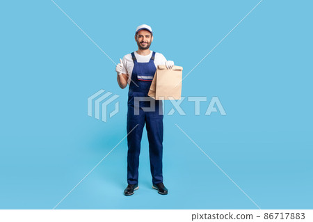 Full length positive courier in blue uniform holding groceries bag and showing thumbs up, like gesture, carrying parcel with goods ordered online. Professional delivery service. studio shot isolated 86717883
