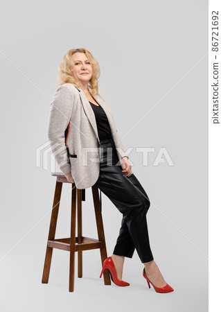 Portrait of senior woman in leather trousers and blazer sitting in profile on over grey studio background 86721692