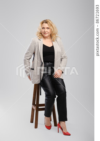 Portrait of senior woman in leather trousers and blazer sitting on tall wooden chair over grey studio background Portrait of senior woman in leather trousers and blazer sitting on tall wooden chair over grey studio background 86721693