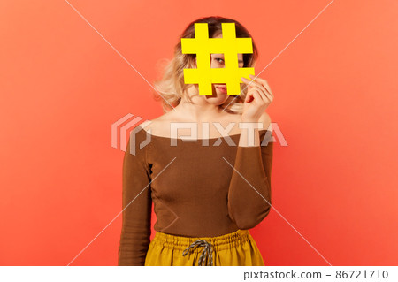 Stylish woman hiding her face behind big yellow hashtag sign, tagging posts in internet, making blogs popular. Indoor studio shot isolated on orange background Stylish woman hiding her face behind big yellow hashtag sign, tagging posts in internet, making blogs popular. Indoor studio shot isolated on orange background 86721710