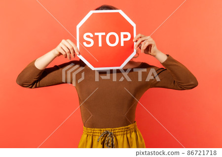 Scared anonymous woman hiding her face behind stop road sign, campaign against domestic violence, protection of women's rights. Indoor studio shot isolated on orange background 86721718