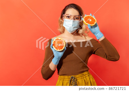 Woman in protective glasses, mask, gloves holding orange fresh fruit, natural vitamins for immune system and flu treatment, healthy nutrition during quarantine. indoor studio shot, red background Woman in protective glasses, mask, gloves holding orange fresh fruit, natural vitamins for immune system and flu treatment, healthy nutrition during quarantine. indoor studio shot, red background 86721721