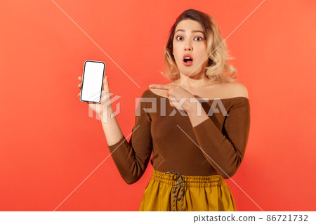 Shocked surprised woman pointing finger at smartphone with white empty screen, looking with amazed expression, online app. Indoor studio shot isolated on orange background Shocked surprised woman pointing finger at smartphone with white empty screen, looking with amazed expression, online app. Indoor studio shot isolated on orange background 86721732