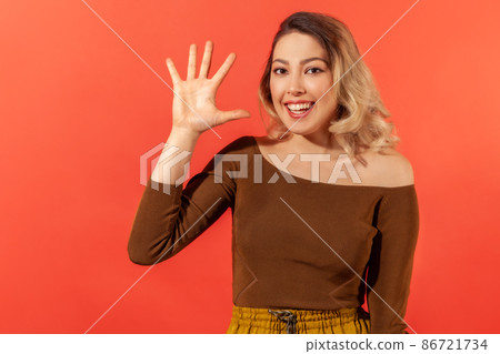 Hello. Portrait of beautiful smiling young lady with blonde hair in brown blouse with raised hand demonstrate hello gesture and ready to make friendship. Indoor studio shoot isolated on red background Hello. Portrait of beautiful smiling young lady with blonde hair in brown blouse with raised hand demonstrate hello gesture and ready to make friendship. Indoor studio shoot isolated on red background 86721734