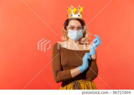 Woman with crown on head, in hygienic mask and protective glasses, adjusting safety gloves to prevent contagious flu coronavirus, 2019-ncov disease. indoor studio shot isolated on red background 86721904