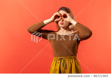Where you. Portrait of serious blonde woman in casual brown blouse making glasses shape, looking through binoculars gesture. Indoor studio shot isolated on red background. Where you. Portrait of serious blonde woman in casual brown blouse making glasses shape, looking through binoculars gesture. Indoor studio shot isolated on red background. 86721905