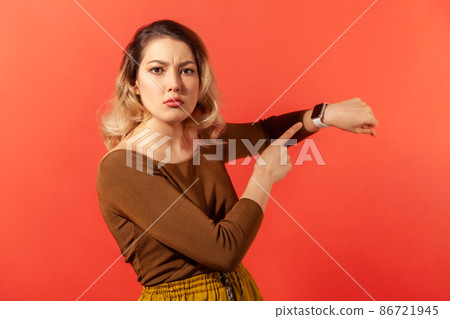 We have no time. Portrait of serious boss woman with blonde hair in brown blouse pointing her finger at the smartwatch and getting angry. Indoor studio shot isolated on red background 86721945