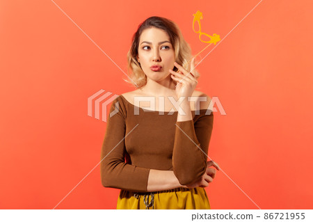 Attractive thoughtful woman holding paper mask on stick contemplating about her costume at fancy party, dreaming. Indoor studio shot isolated on orange background Attractive thoughtful woman holding paper mask on stick contemplating about her costume at fancy party, dreaming. Indoor studio shot isolated on orange background 86721955