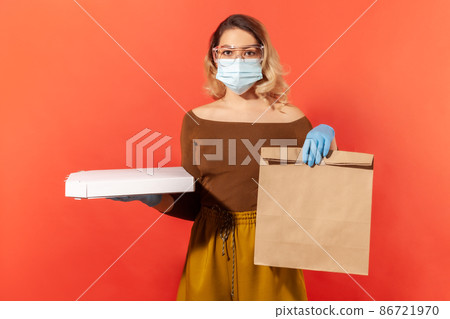 Safe food shopping on quarantine. Woman in protective mask, gloves, glasses holding groceries bag and pizza, delivery service in coronavirus self-isolation, online order. indoor studio shot, isolated Safe food shopping on quarantine. Woman in protective mask, gloves, glasses holding groceries bag and pizza, delivery service in coronavirus self-isolation, online order. indoor studio shot, isolated 86721970