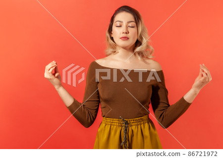 Portrait of beautiful calm young woman with blonde hair in brown blouse standing with closed eyes, meditation with mudra gesture, peaceful mind. Indoor studio shot isolated on red background Portrait of beautiful calm young woman with blonde hair in brown blouse standing with closed eyes, meditation with mudra gesture, peaceful mind. Indoor studio shot isolated on red background 86721972