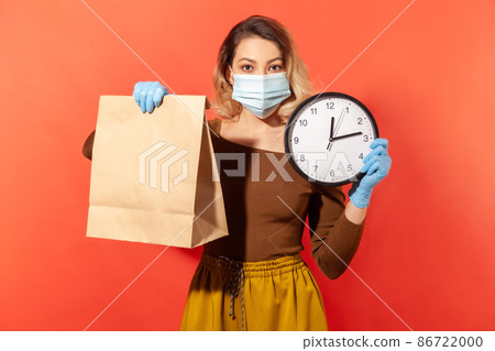 Time to order food, safe shopping on quarantine. Woman in protective mask holding paper bag and big clock, delivery in coronavirus self-isolation. indoor studio shoot isolated on red background 86722000