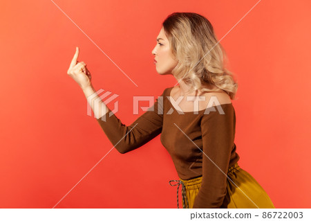 Side view portrait of angry serious young woman with blonde hair in brown casual blouse standing and shows fuck sign. Indoor studio shot isolated on red background 86722003