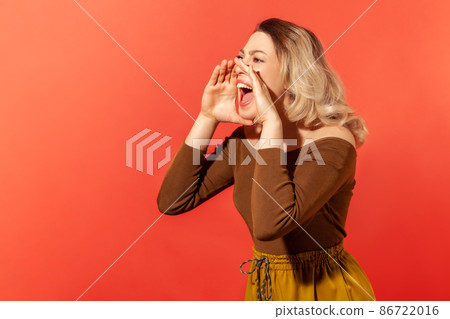 Portrait of angry woman with blonde hair in brown blouse standing and screaming about bad news or troubles holding her hands near mouth. Indoor studio shot isolated on red background Portrait of angry woman with blonde hair in brown blouse standing and screaming about bad news or troubles holding her hands near mouth. Indoor studio shot isolated on red background 86722016