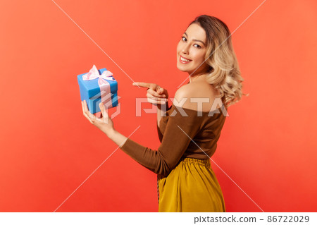 Side view beautiful woman pointing finger at little blue giftbox with pink ribbon, presents and bonuses. Indoor studio shot isolated on orange background Side view beautiful woman pointing finger at little blue giftbox with pink ribbon, presents and bonuses. Indoor studio shot isolated on orange background 86722029