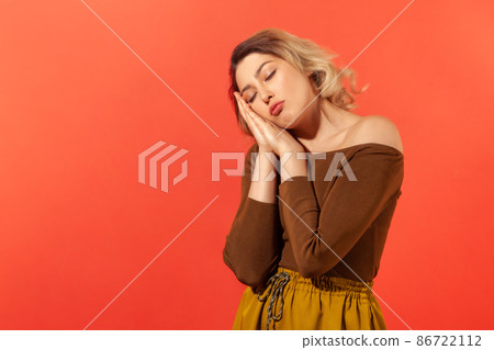I want to sleep. Portrait of woman with blonde curly hair with closed eyes standing with arms under her head like pillow and sleep. Indoor studio shot isolated on red background I want to sleep. Portrait of woman with blonde curly hair with closed eyes standing with arms under her head like pillow and sleep. Indoor studio shot isolated on red background 86722112