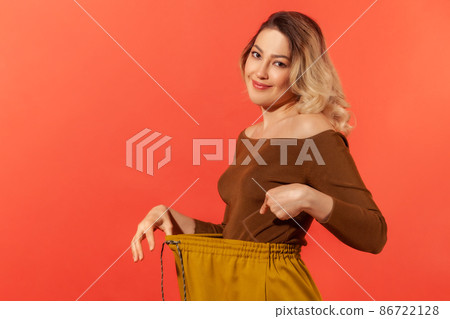 Portrait of beautiful young adult woman have willpower showing slim waist in big trouses, successful weight loss, diet conception. Indoor studio shot isolated on red background Portrait of beautiful young adult woman have willpower showing slim waist in big trouses, successful weight loss, diet conception. Indoor studio shot isolated on red background 86722128