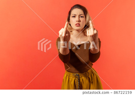 Portrait of angry serious young woman with blonde hair in brown casual blouse standing and shows fuck sign with both hands into the camera. Indoor studio shot isolated on red background 86722159