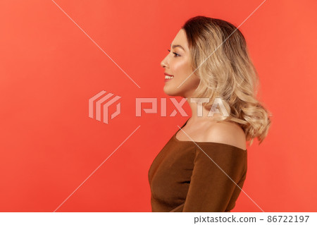 Side view portrait of pretty girl with blonde hair in brown blouse, she looks to the side, smiles, express happiness, energy and friendliness. Indoor studio shot isolated on red background 86722197