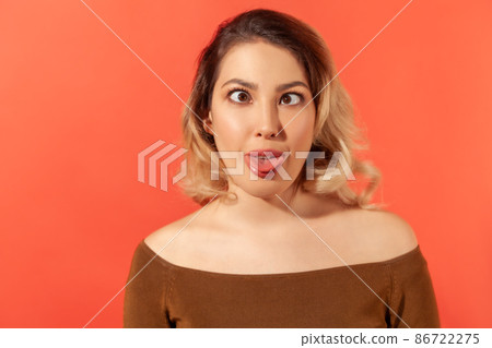 Portrait of funny crazy young beautiful blonde woman in brown blouse standing and looking with crossed eyes and tongue out. Indoor studio shot isolated on red background 86722275