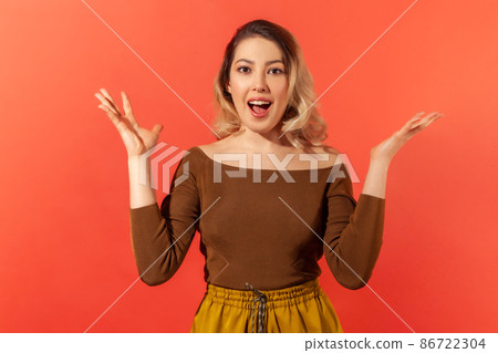Portrait of beautiful amazed pretty woman with blonde hair in brown blouse standing surprised with open mouth, raised hands and looking at the camera. Indoor studio shot isolated on red background 86722304