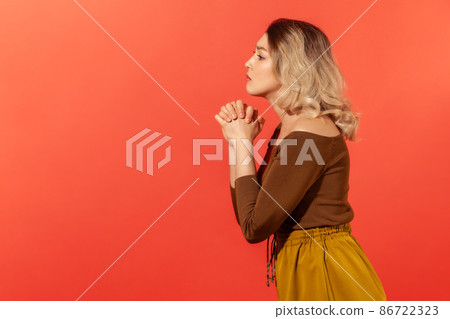 Side view portrait of upset blonde woman standing and took hands together and pray, asking to help her. Worry about problems in life. Indoor studio shoot isolated on red background 86722323