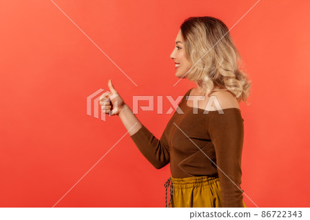 Side view portrait of happy woman with blonde hair and toothy smile, she looks satisfied and positive, demonstrate it by do thumb up gesture. Looking to the side. Indoor isolated on red background Side view portrait of happy woman with blonde hair and toothy smile, she looks satisfied and positive, demonstrate it by do thumb up gesture. Looking to the side. Indoor isolated on red background 86722343