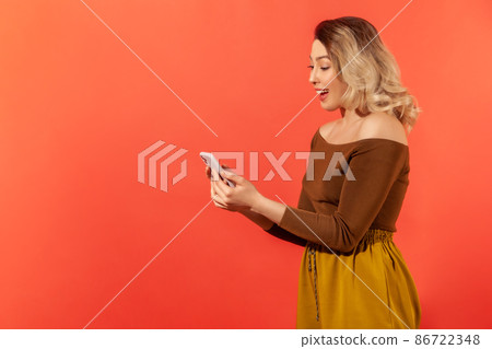 Side view happy surprised woman holding smartphone, reading pleasant message or email and smiling. Indoor studio shot isolated on orange background 86722348