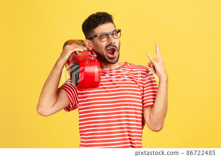 Extremely happy joyful man with beard in eyeglasses and red striped t-shirt holding tape recorder on shoulder and showing rock and roll gesture. Indoor studio shot isolated on yellow background 86722485