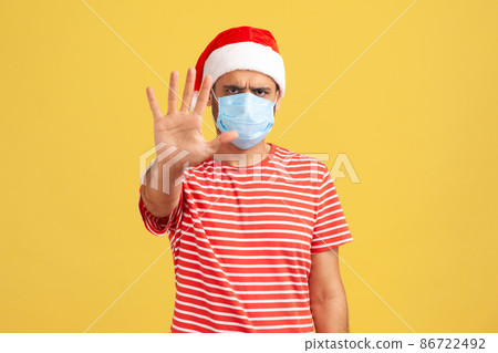 Don't come to me and keep distance. Concerned man in medical face mask and santa claus hat holding hand showing stop gesture. Indoor studio shot isolated on yellow background 86722492