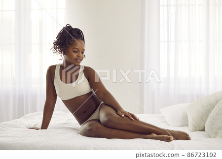 Beautiful young African American woman in underwear sit relax on bed at home. Happy pretty confident black ethnic female in lingerie feel body positive about natural curvy figure shape. 86723102