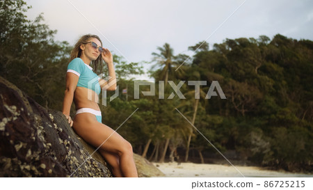 Woman leaning on a rock reef enjoying and relax warm dark golden sunset. Palm tree in beach. Woman in blue swimsuit and sunglasses 86725215